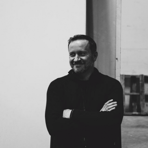 waste-up photo of Region 6 installation artist James Culleton with short, dark hair, wearing a long-sleeve black shirt, crossing his arms, looking left and closed smile