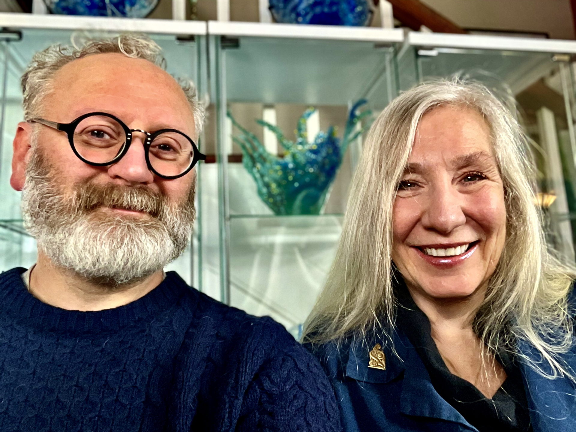 head and shoulders of brightly smiling Alan and Nicole Milligan with fair skin, grey hair, grey beard, and black round rimmed glasses on Alan