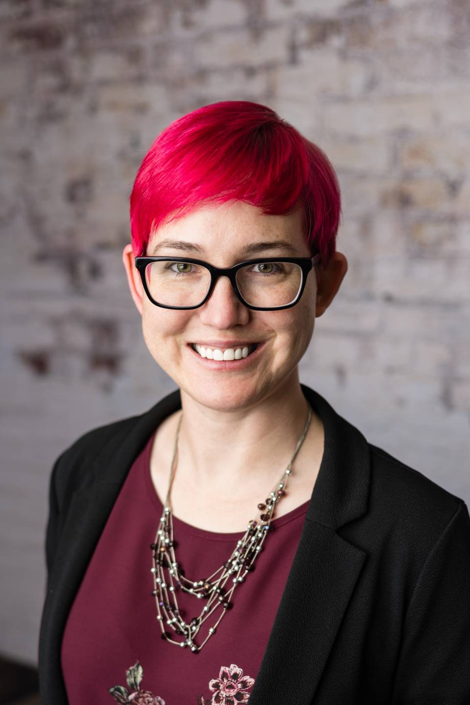 Chest-up image of caucasian Amanda Skinner with hot pink hair, dark-rimmed glasses, wearing a black suit jacket with a burgundy under shirt, a black beaded necklace and warm toothy smile