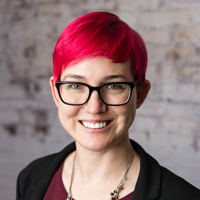 Chest-up image of caucasian Amanda Skinner with hot pink hair, dark-rimmed glasses, wearing a black suit jacket with a burgundy under shirt, a black beaded necklace and warm toothy smile