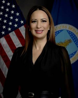 Waste-up image of Kristy Zaste wearing a v-neck, long-sleeve black shirt with light brown long hair, dark eyes, and slightly toothy smile, with American and North Dakota flags behind her