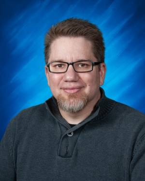 Formal chest-up image of Derek Morin wearing a collared dark-grey sweater with a full-head of dark hair, salt and pepper goatee, dark-rimmed rectangular glasses, light skin and a closed smile