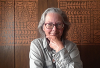 Bowman artist Cris Fulton with shoulder-length grey hair, wearing dark-rimmed glasses, a long-sleeve blue jean shirt, and a closed smile that reaches her eyes