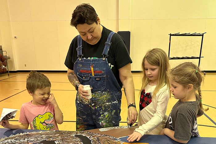 Teaching artist Anna Johnson, wearing a messy apron, painting on a canvas with 3 Wyndmere Elementary students