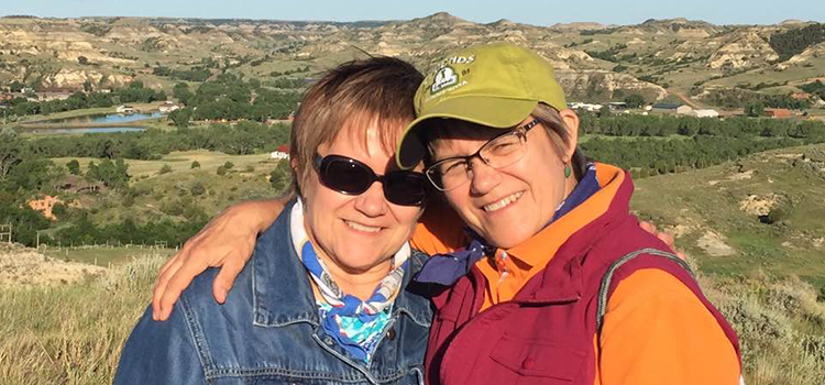 Barbara and Beverly Benda with short brown hair, looking into the sun at the camera with the North Dakota badlands in the background on a cool summer day