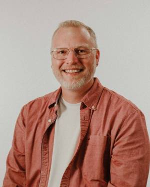 Head and shoulders of Matthew Anderson wearing a dark coral button-up shirt with a white undershirt, clear rimmed glasses, with pale skin, a big toothy smile, dark eyes, short, light hair and short gray beard