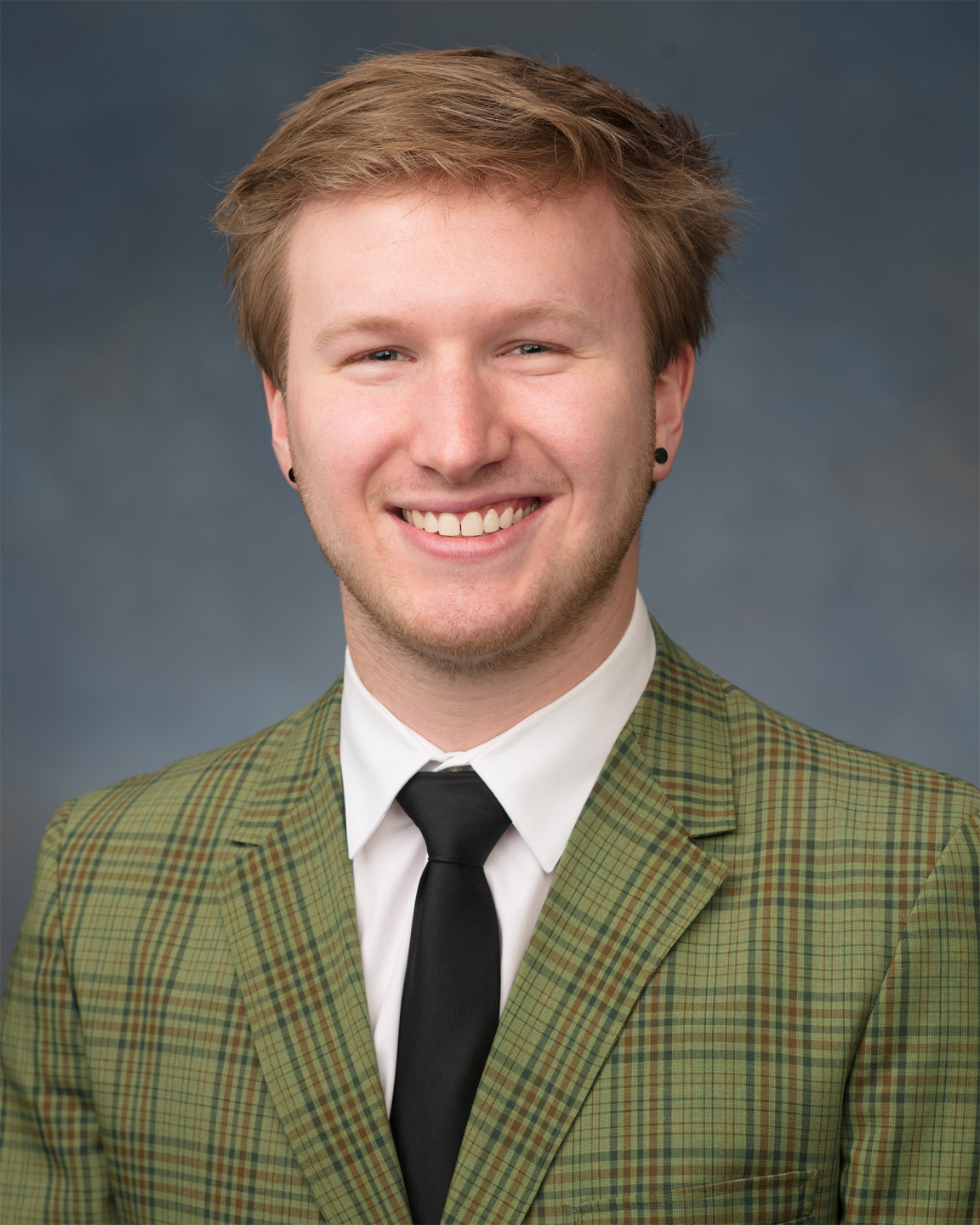 head and shoulders of Tobias Zikmund with short, messy, dirty blonde hair, black dot earrings, pale skin and a big toothy smile; wearing a green striped suit jacket and black tie