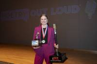 2025 ND POL Champion Leah Hochhalter of Valley City H.S., smiling brightly, wearing a magenta pants suit, standing on a stage, holding a large trophy in one hand and smaller trophy in another