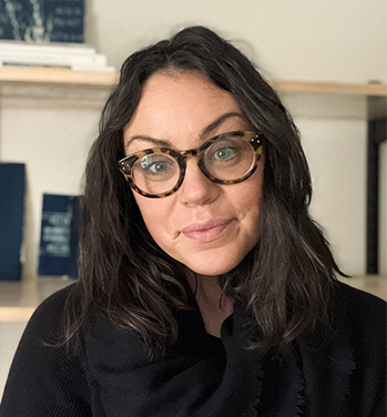 head and shoulders of Jessica Christy wearing a black shirt, thick, dark patterned glasses, long, thick, straight dark hair, pale skin, grey/blue eyes and a sweet closed smile