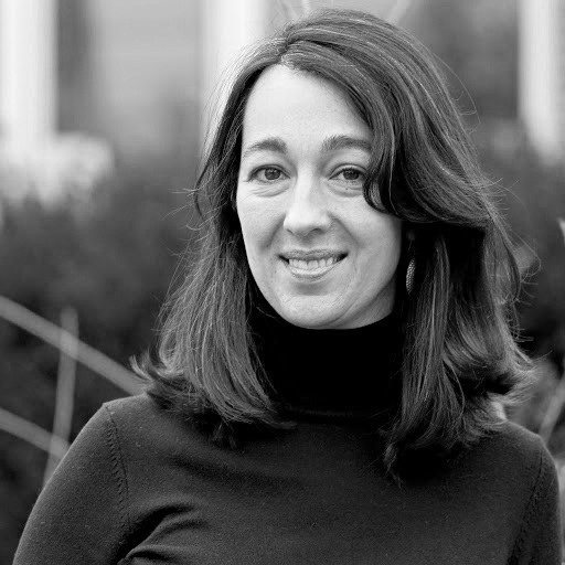 Greyscale - head and shoulders of Teva Dawson with pale skin, shoulder-length, thick, wavy, dark hair with strands of grey and dark eyes; wearing a dark turtle-neck sweater and sweet toothy smile with a blurred outdoor background