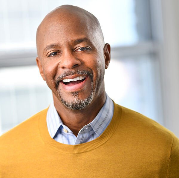 Head and shoulders of African American Torrie Allen, with a bald head, salt and pepper goatee, wearing a mustard-colored sweater and a big toothy smile