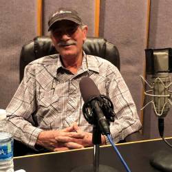 Frank Kuntz, sitting in front of a microphone on a table, wearing a button-down cotton shirt and baseball cap; with grey hair and mustache