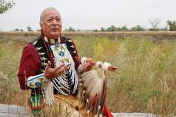 Native American storyteller Keith Bear, wearing colorful Native clothing and using his hands to tell a story while sitting on a bench with tall grass all around him