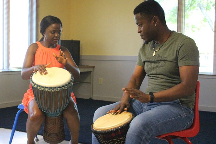 Hamzat, seated on the right, leads a drumming session with a woman in an orange dress. Both are playing traditional hand drums in a brightly lit room, focused on rhythm and technique