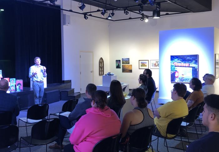 A man stands and speaks to a group of people sitting in an art gallery.