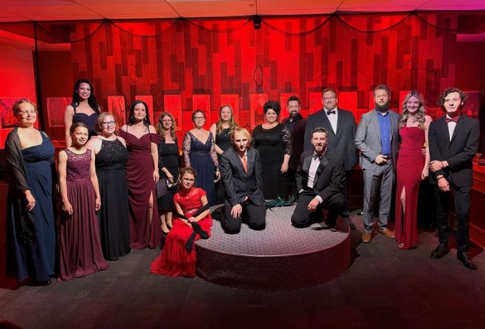 A group of people in dresses and suits stands in front of a red curtain.