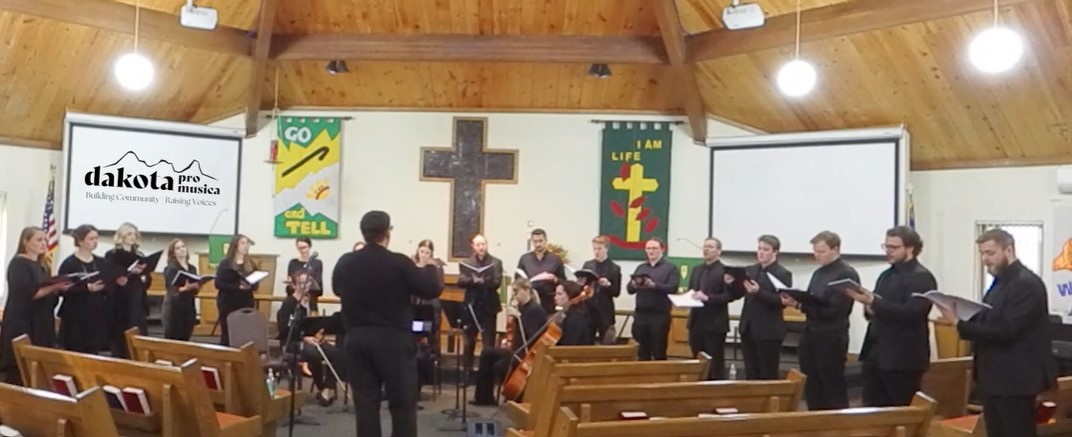 A male conductor in front of a line of people singing, holding songbooks.