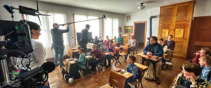 A film crew and children in desks in an old schoolhouse.