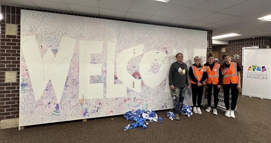 A large sign with 'Welcome' on it in white surrounded by colors, four people stand in front of sign.