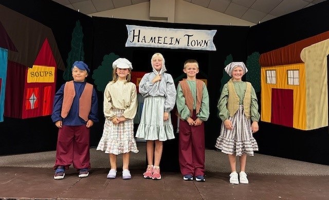 Three girls and two boys stand on stage in dresses, vests, and hats..