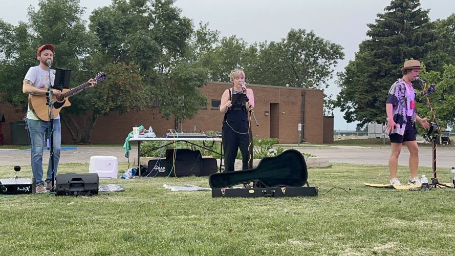 Man playing guitar, woman singing, man singing on lawn in front of brick building.