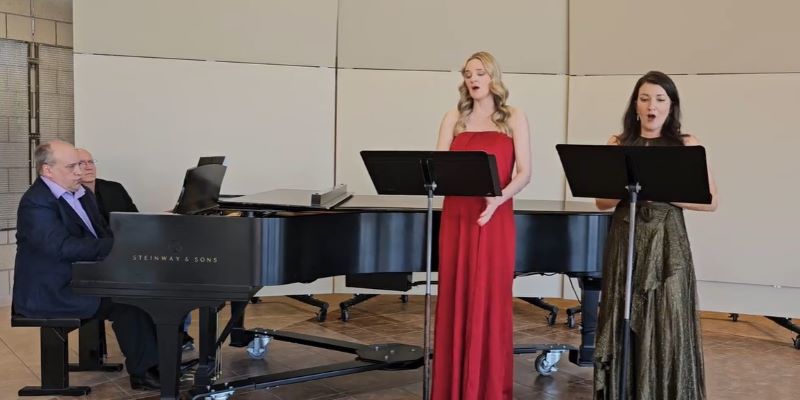 A man plays piano, a man sits beside him, a woman in a red dress sings, a woman in an olive green dress sings.