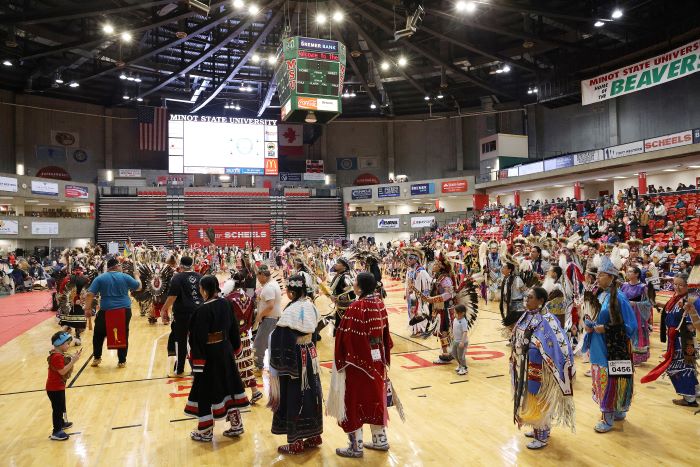 People in colorful Native American dress walk and dance in a gym.