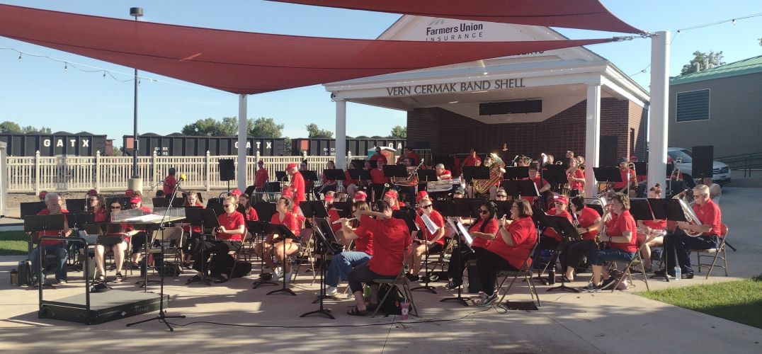 Many people sit in front of a band shell playing instruments on a summer evening.