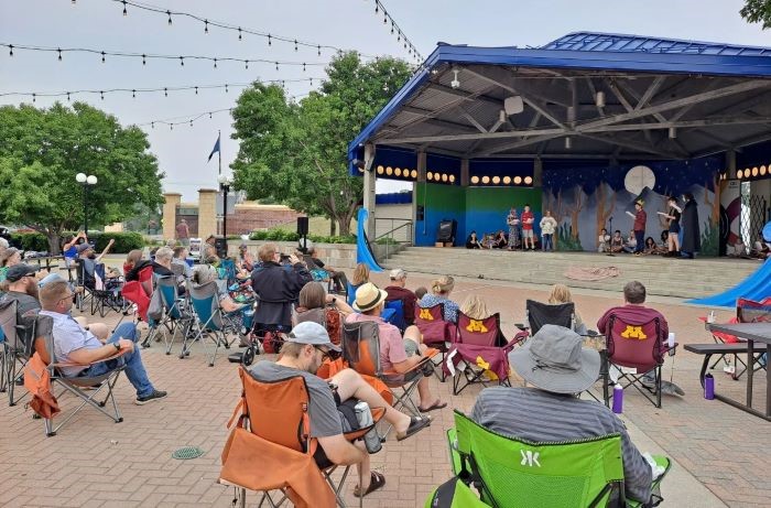 People sit in lawn chairs on a patio while actors perform on a stage.