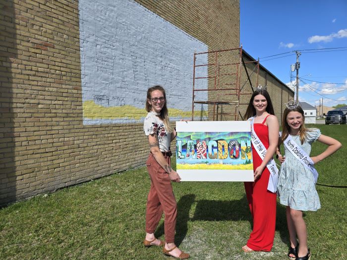 Two women hold a painting, a girl stands beside them. A mural is started on a brick wall.