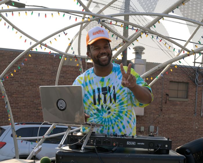 A smiling man gives a peace sign as he deejays with musical equipment.