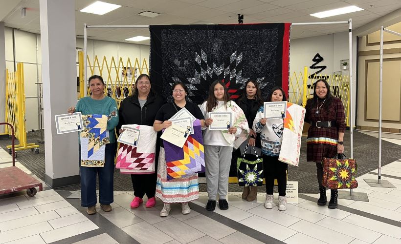 Seven women stand holding certificates in front of a black quilt.