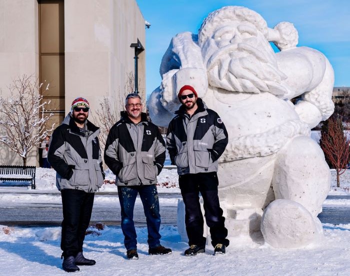 Three men in gray and black coats stand next to a snow sculpture of Santa Claus.