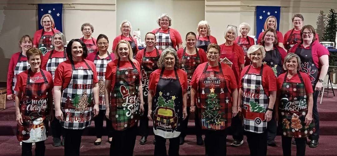 Three rows of women stand smiling, wearing red shirts and Christmas-themed aprons.