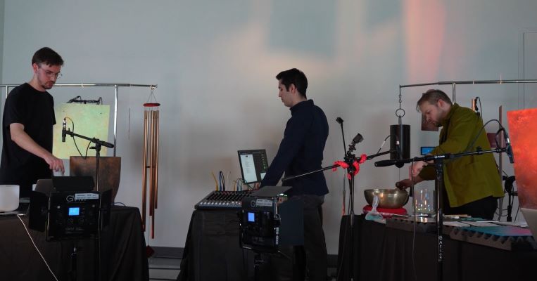 Three men play music, one on synthesizer, one on computer, one playing bowl.