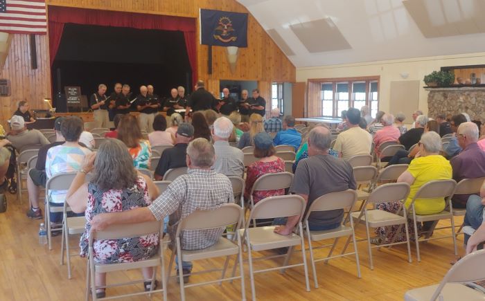 People sit in chairs and listen to a group of men in black shirts singing.