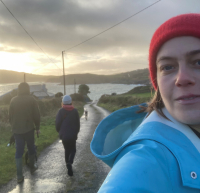 Artist Mollie Douthit taking a selfie in Ireland with people walking behind her on a narrow paved street that leads to a small lake, on a lightly raining day with the sun peaking over the horizon