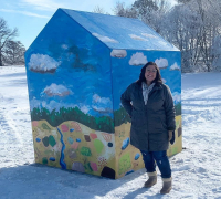 Nicole Gagner standing outside on snow-covered ground in front of a 12x12 foot colorfully painted with scenic elements - house on a sunny day with frosted trees behind her