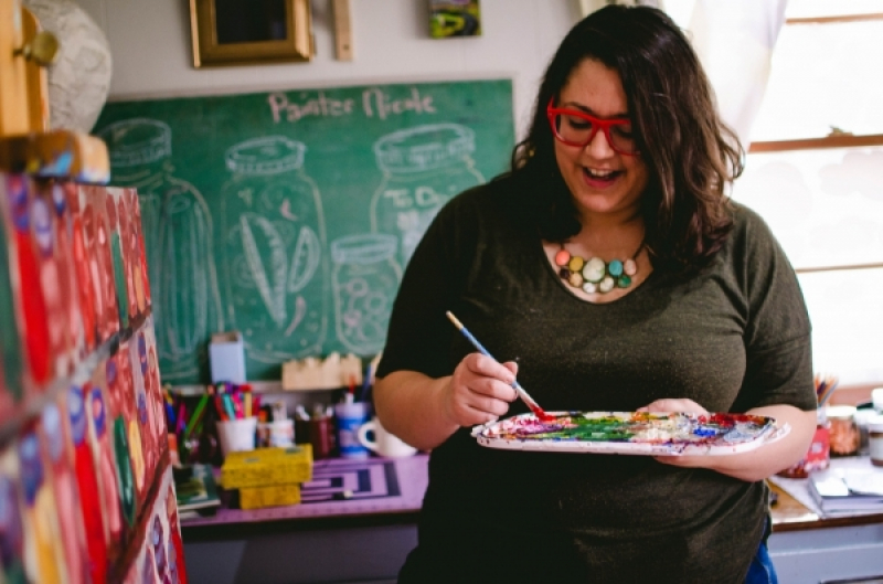 Teaching artist Nicole Gagner mixing paint, wearing red-rimmed glasses, with long, thick wavy dark hair, enjoying herself