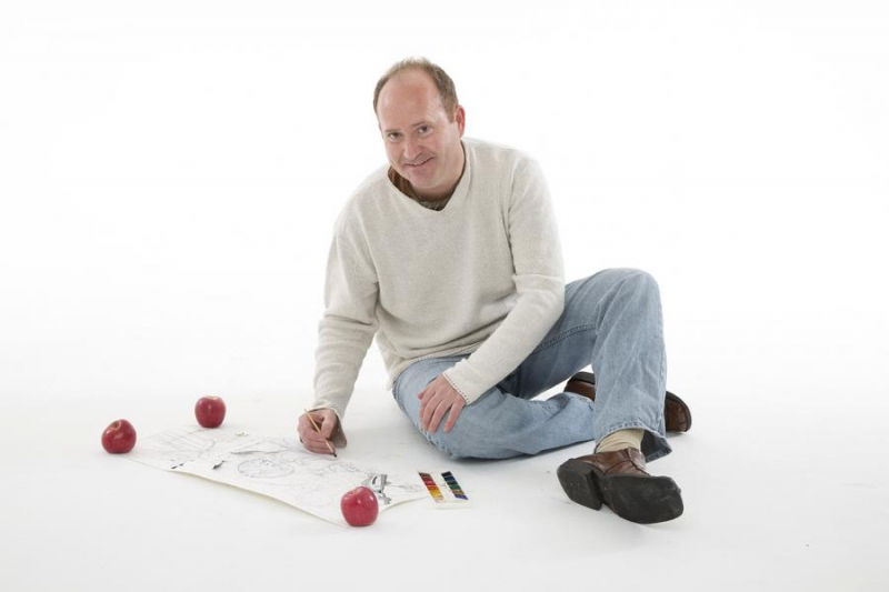 Teaching artist Markus Tracy with balding brown hair, smiling brightly, sitting on a white floor with crossed legs, wearing blue jeans and a white sweater, playing a game with red balls
