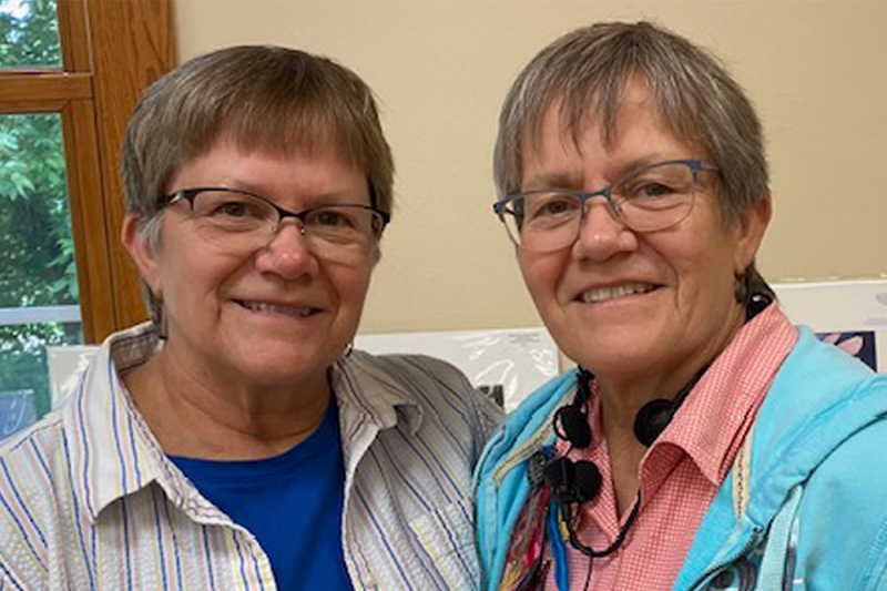Head and shoulders of sisters Barbara and Beverly Benda, with short brown/grey hair, matching cat-style glasses and toothy smiles